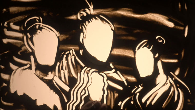 Three figures drawn out of sand on a lightbox, the figures are wearing cloaks and have top knots.