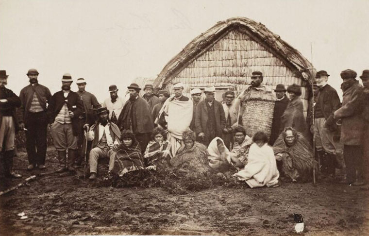 Group of 25 people infront of a flax house, the people are wearing a combination of european clothes and traditional Māori cloaks.