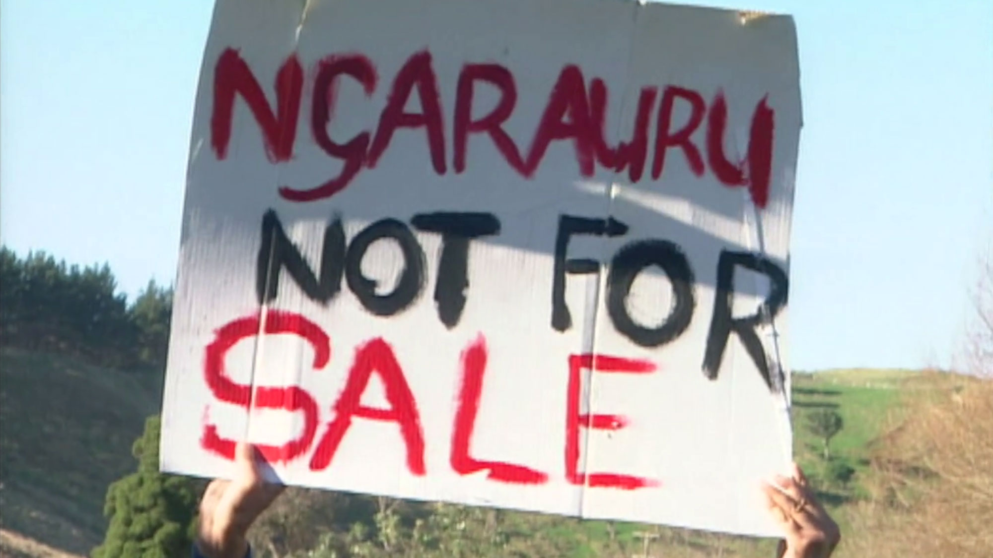 Hands holding up a hand-painted sign reading ' NGA RAURU NOT FOR SALE'