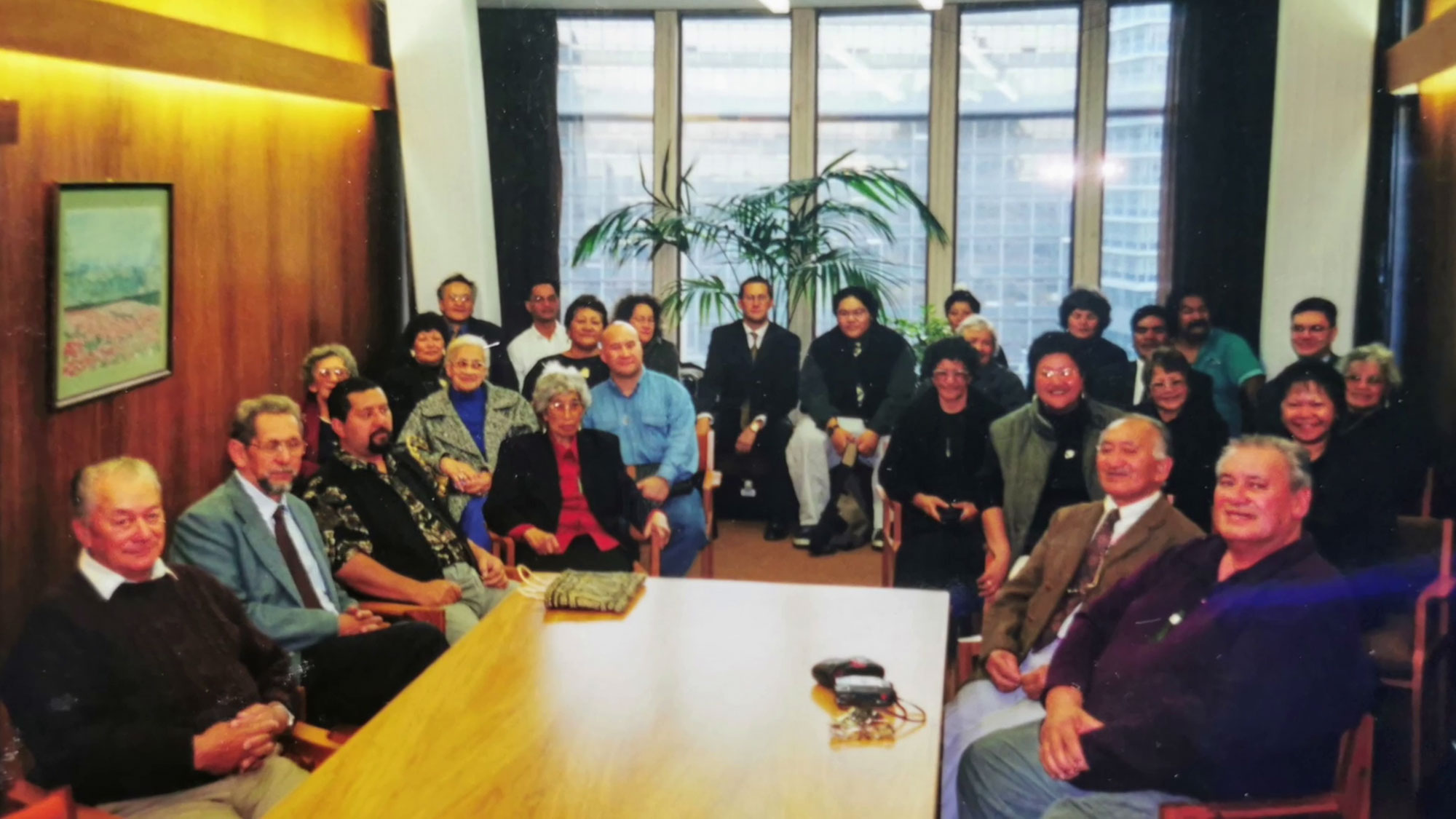 A group of people seated in a meeting room with large windows, a wooden wall and wood table.