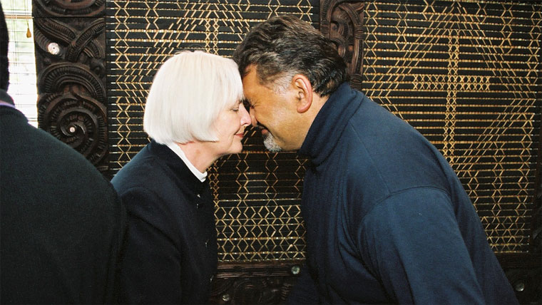 A woman with white hair giving a man a hongi in front of tukutuku panels.