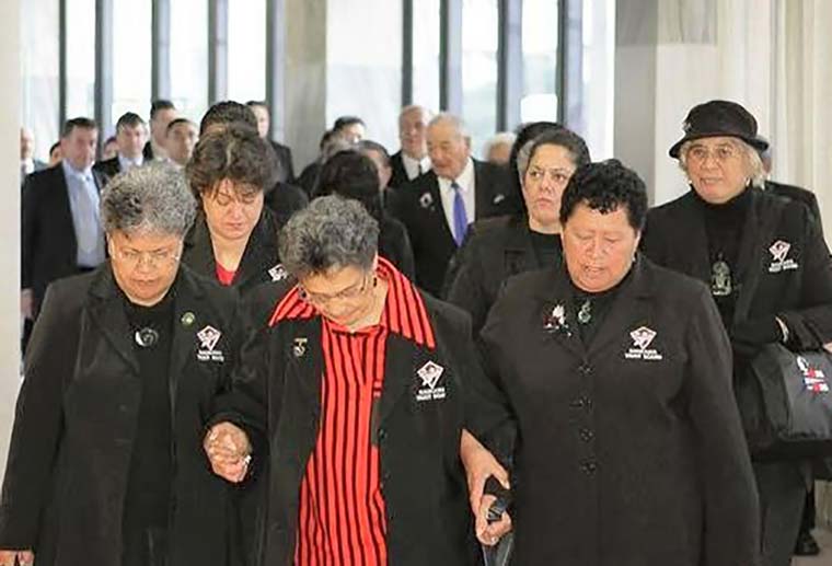 Women with heads bowed holding hands in foreground with men behind.