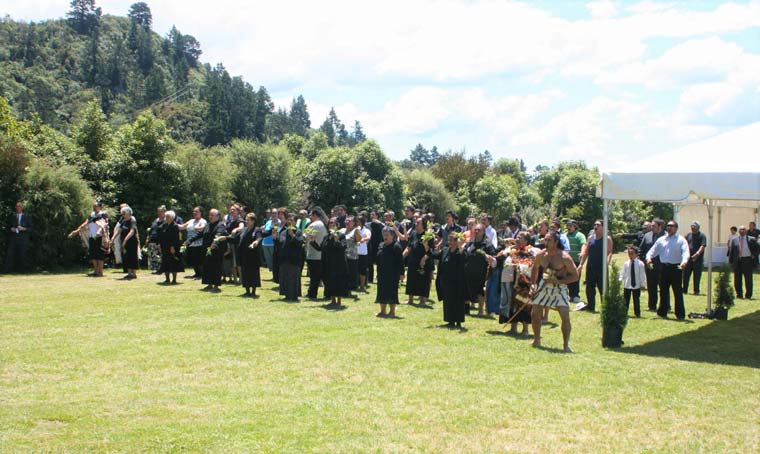 Women in black with leaves singing and dancing with a bare-chested man with a taiaha to their right. Set on a lawn of grass near a temporary canvas shelter.