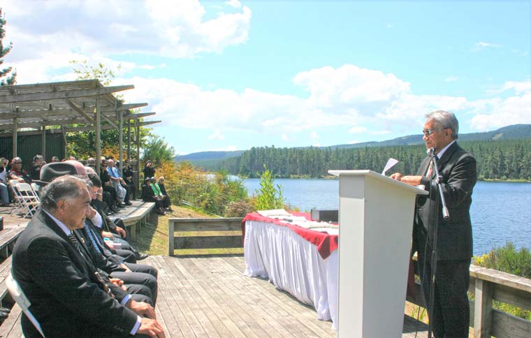 Mana at a lectern speaking to seated audience. Located on the banks of a river'.