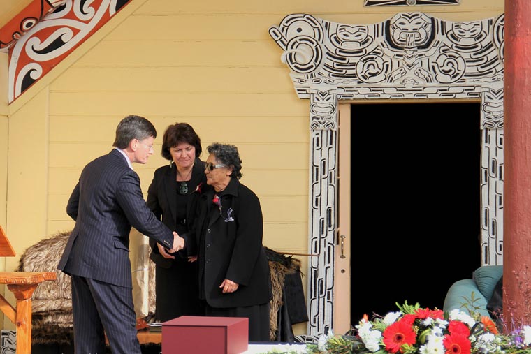 Minister shaking hand of kuia in front of a whare. Another woman stands nearby.