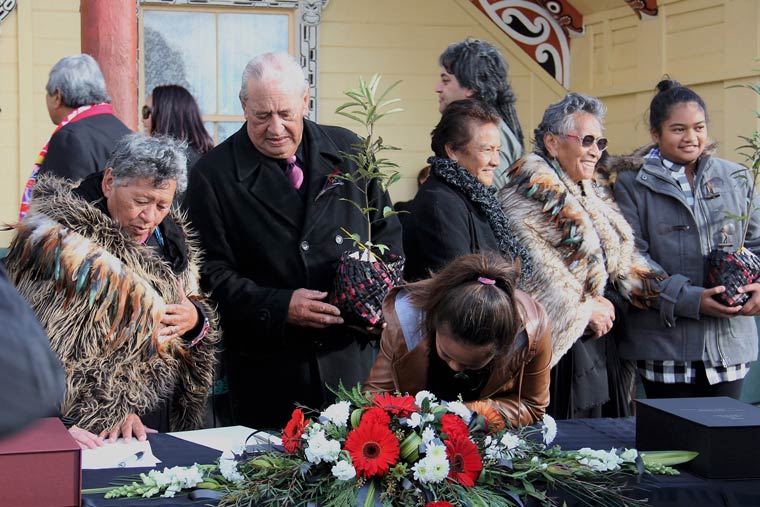 Group gathered a woman signing a document on a table covered in a wreath of flowers.