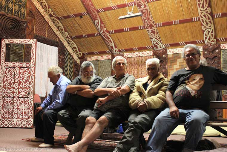 Five kaumatua sitting on bench inside a marae building.