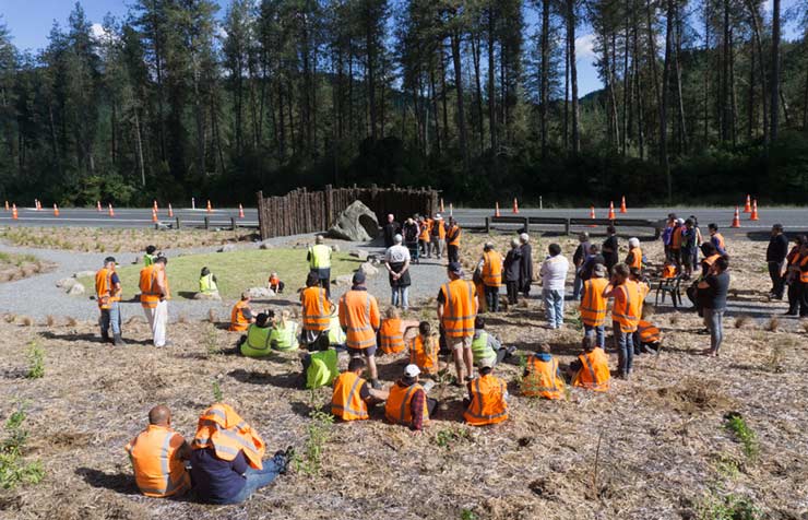 Group in orange vests listening to a speaker standing in front of a stone memorial next to the road