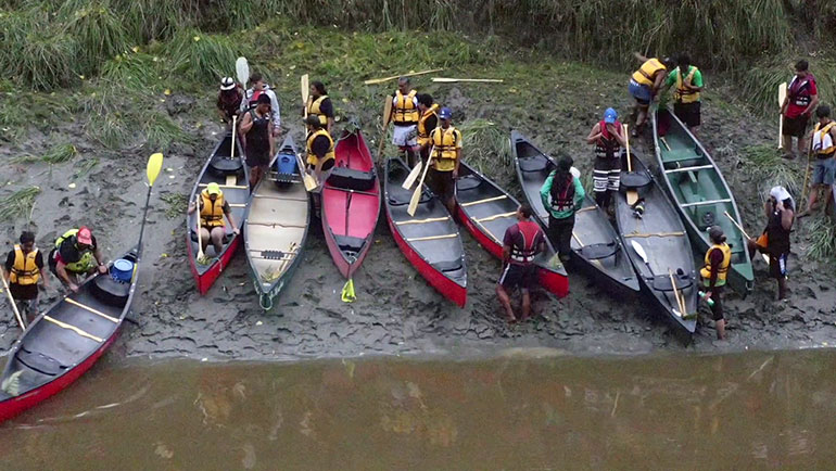 Group of kayaks and kayakers gathered on side of a river