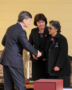 Minister shaking hand of kuia in front of a whare. Another woman stands nearby.